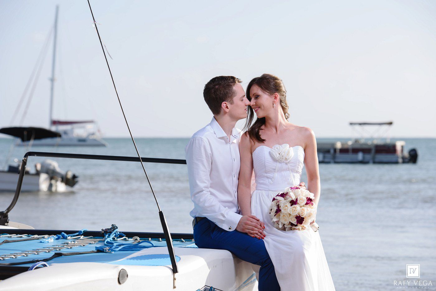 Newlywed couple sharing a romantic moment on a sailboat by the sea, bride holding a bouquet in a white gown.