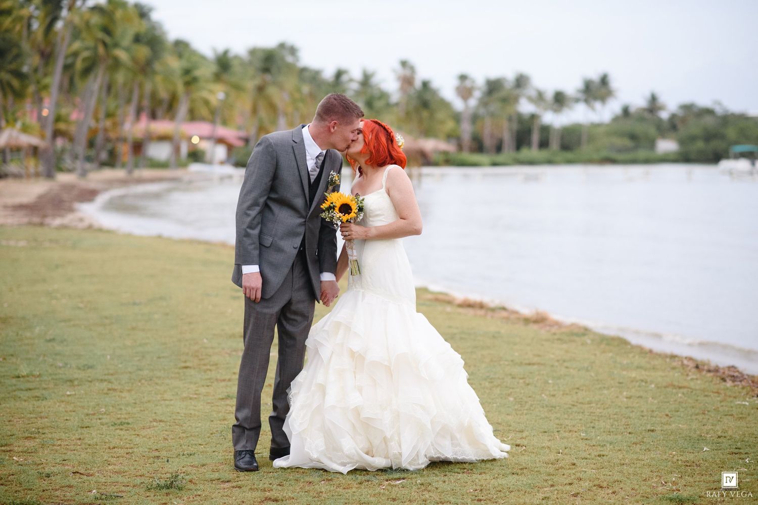 Bride in white gown with sunflower bouquet and groom in gray suit share a romantic moment by a tropical beach.