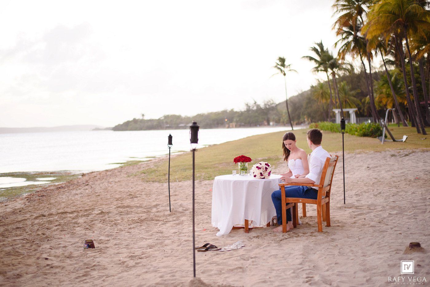Couple enjoying a romantic beachside dinner at sunset with a white tablecloth, red roses, and palm trees in the background.