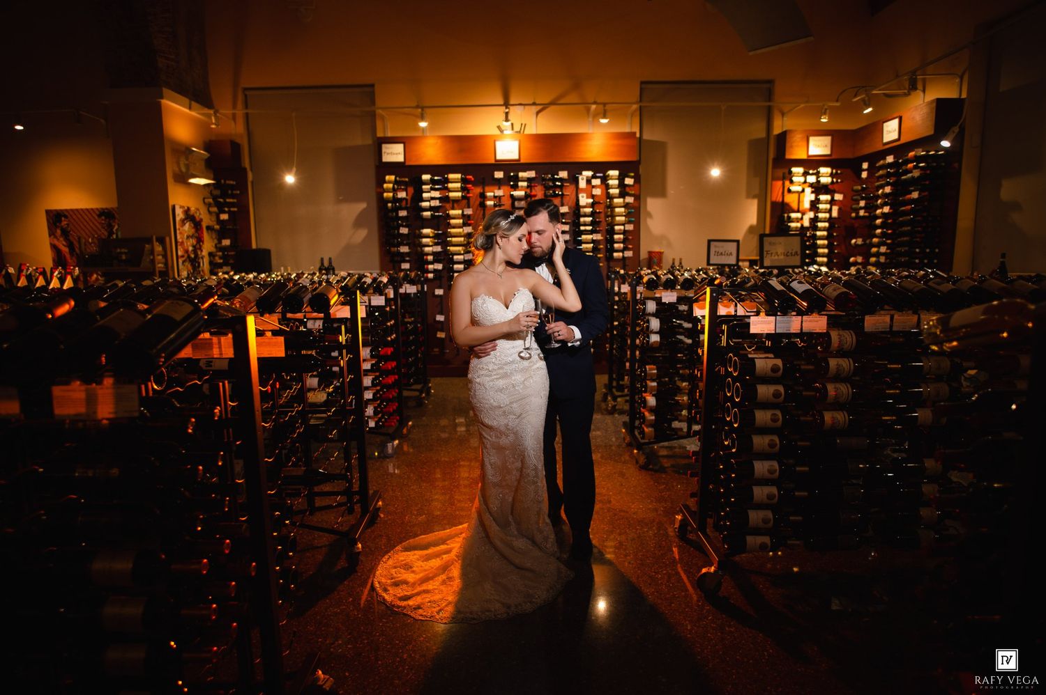 Bride and groom embrace romantically in a warmly lit wine cellar surrounded by floor-to-ceiling wine racks on their wedding day.