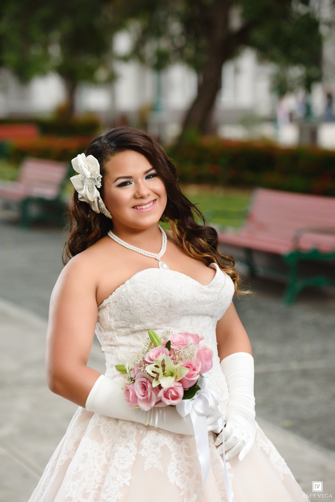 Young woman in white strapless lace gown, long gloves, pearl necklace, and floral hair accessory holds pink rose bouquet.