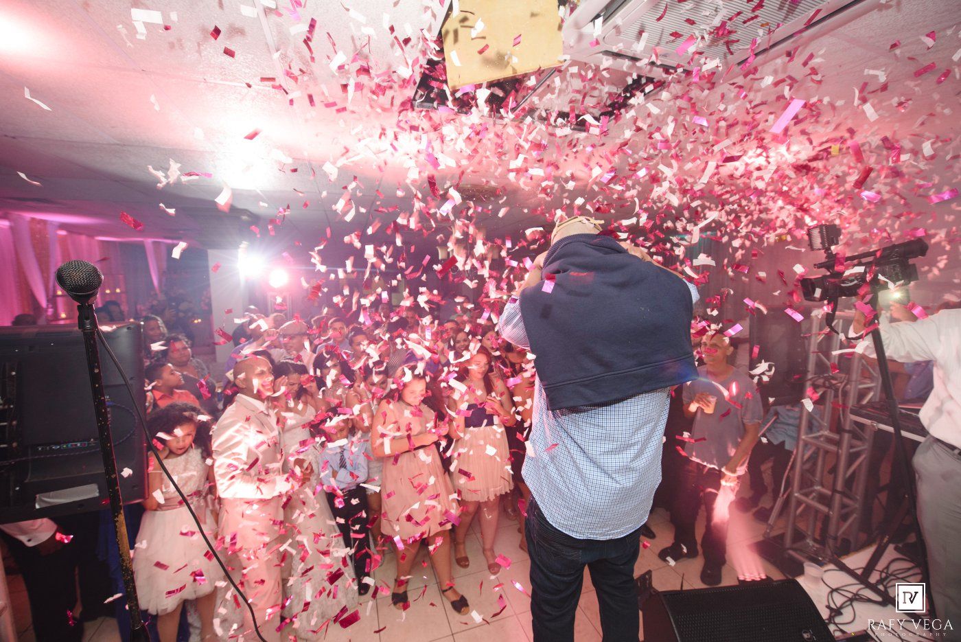 Confetti explosion fills a lively party venue as a performer entertains a crowd of guests under pink lighting.