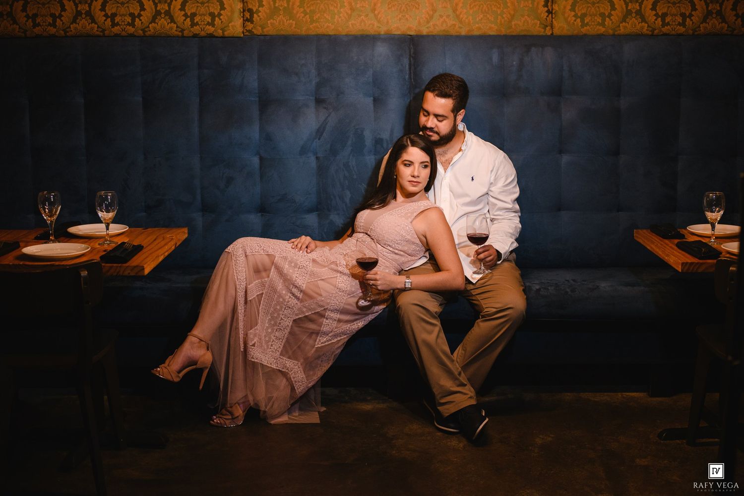 Couple posing romantically on a blue velvet booth in a dimly lit restaurant, holding wine glasses.