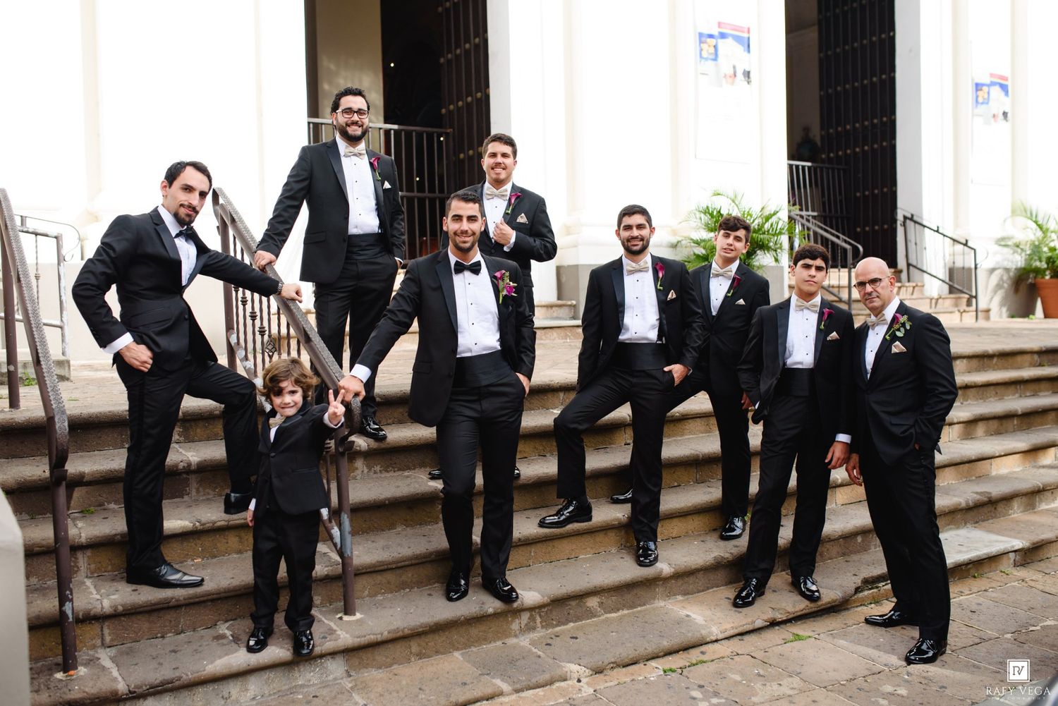 Groomsmen in black tuxedos posing on church steps with a young ring bearer at a formal wedding.