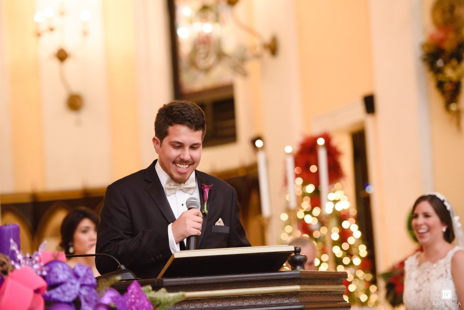 A smiling groom in a tuxedo speaks into a microphone at a podium during a festive wedding reception with Christmas decorations.
