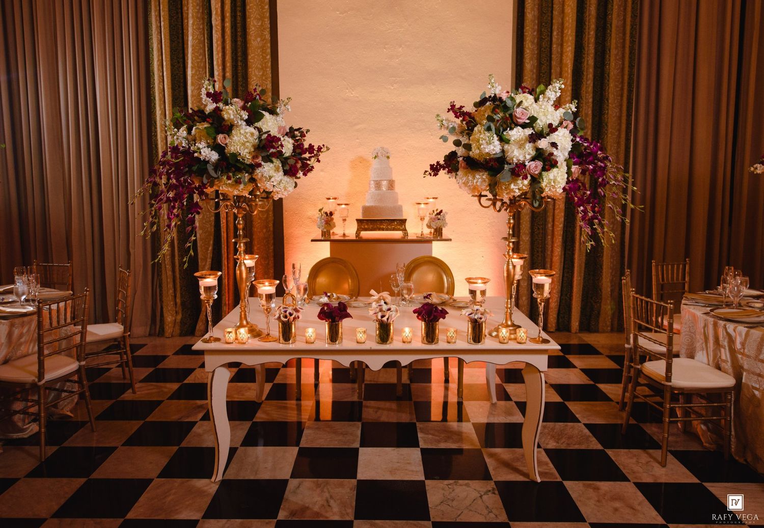 Elegant wedding reception table with floral arrangements, candles, and a tiered cake on a black and white checkered floor.