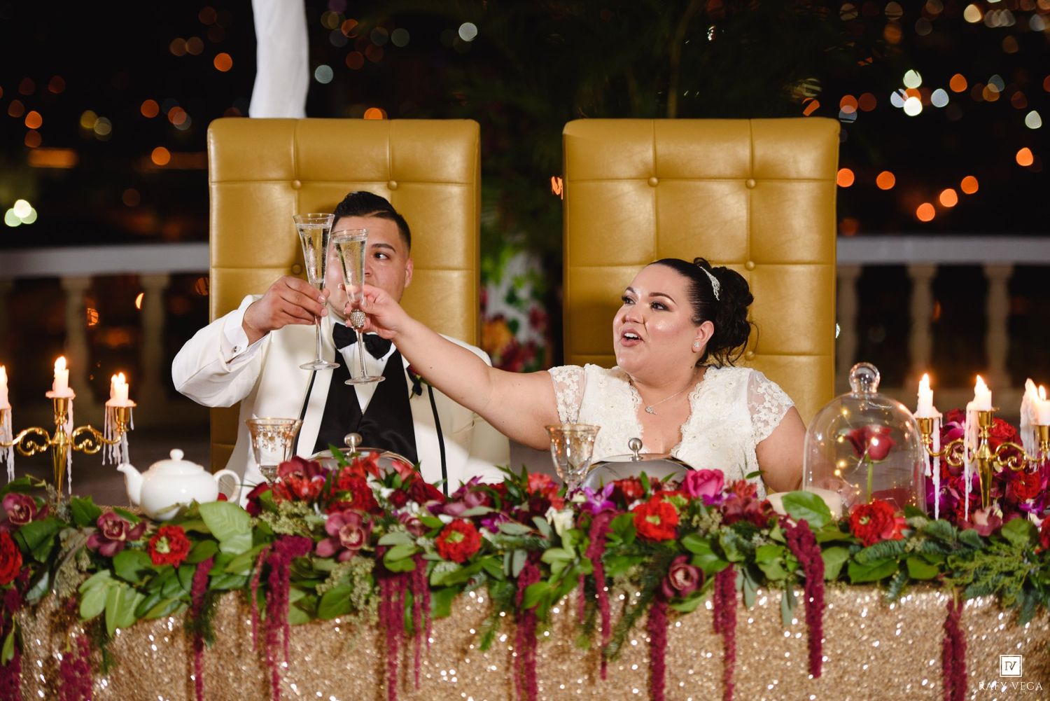 Bride and groom toasting champagne glasses at elegant wedding reception table adorned with red floral arrangements and candles.