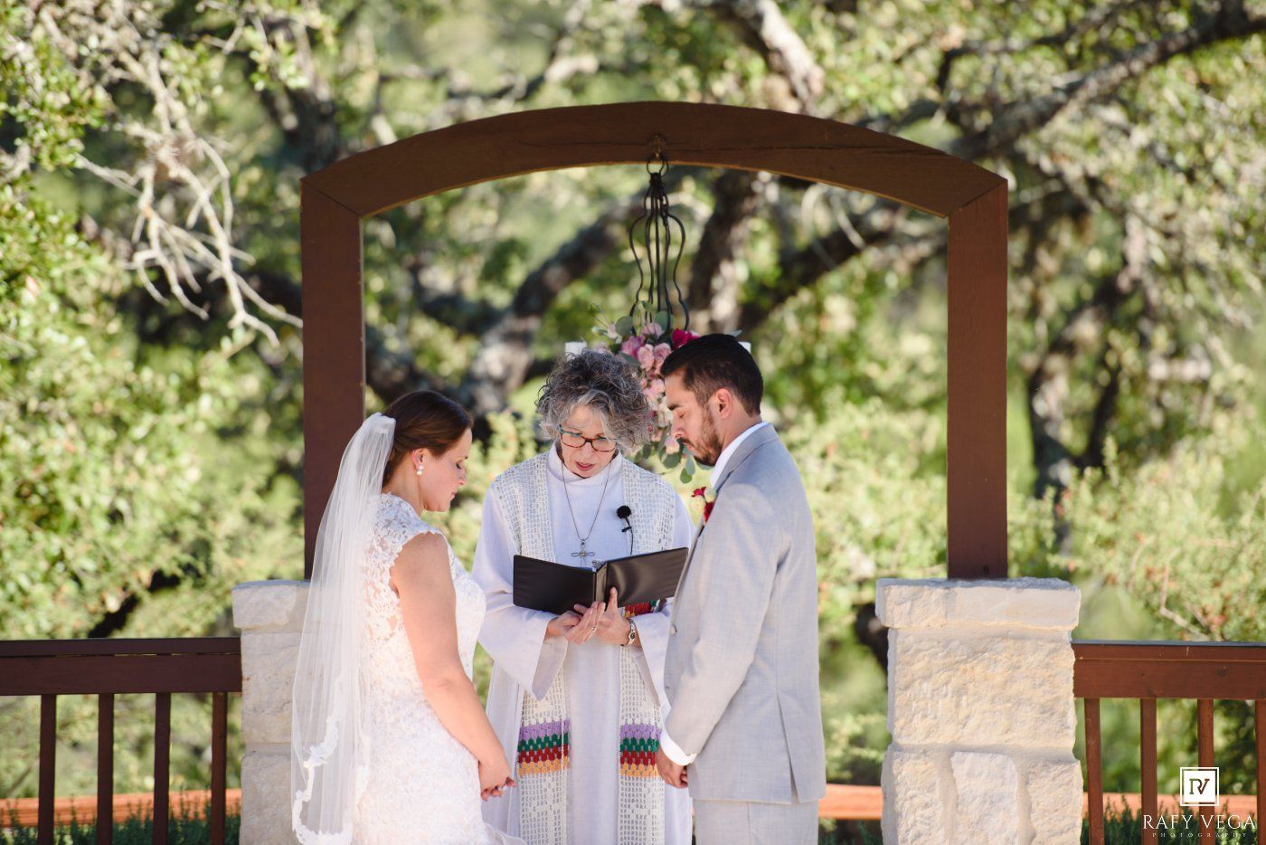 Bride and groom exchange vows with officiant under wooden arch at outdoor wedding ceremony surrounded by lush green trees.