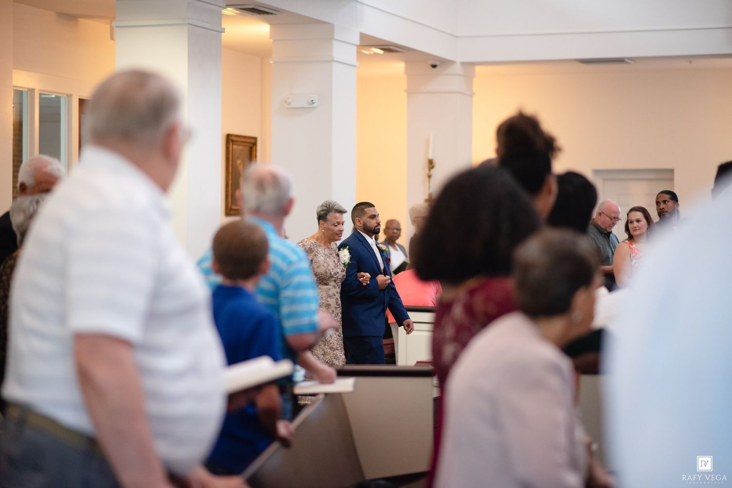 Congregation standing during a church service with a man in a blue suit speaking at the front.