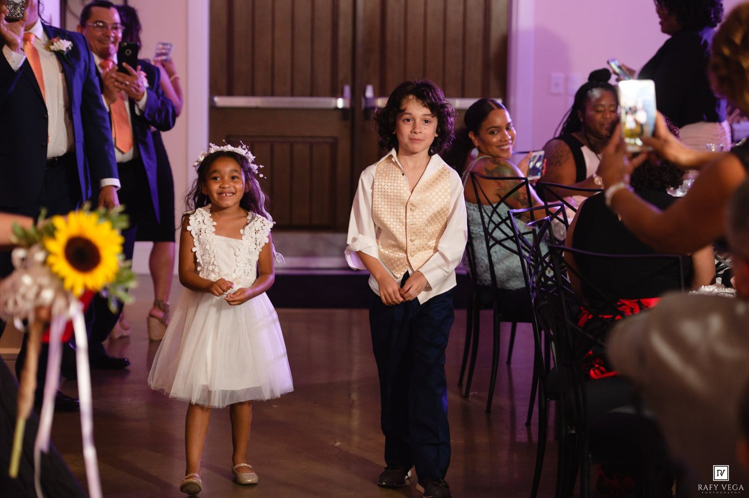 Two children dressed formally walk down an aisle at a wedding reception in an elegant venue.