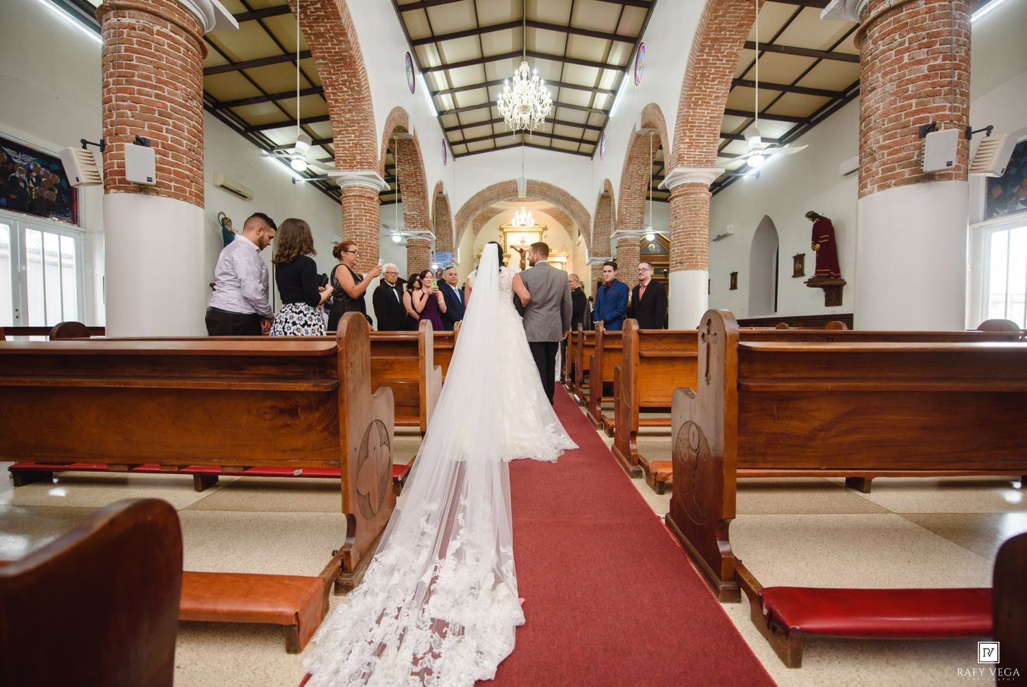 Bride in elegant white gown with long veil walks down red carpet aisle in historic brick church during wedding ceremony.