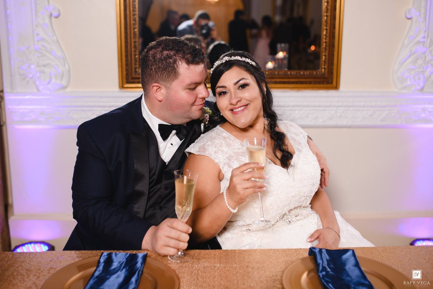 Bride and groom toasting with champagne glasses at elegant wedding reception, groom in tuxedo kissing bride's cheek.