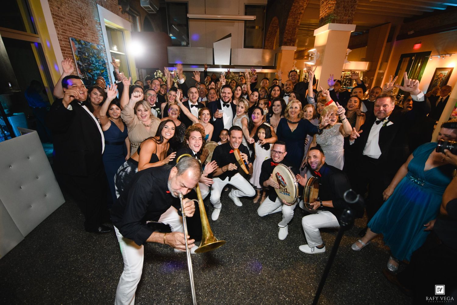 Large group of elegantly dressed guests posing with a live band at a festive indoor event celebration.