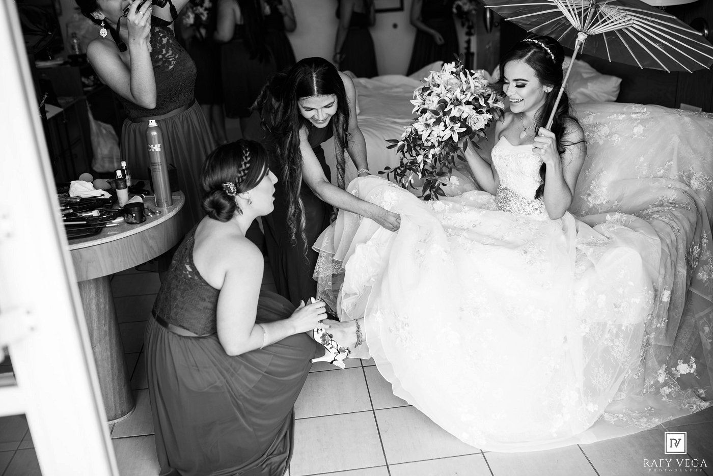 Bridesmaids help bride in white gown prepare for wedding ceremony in black and white photo.
