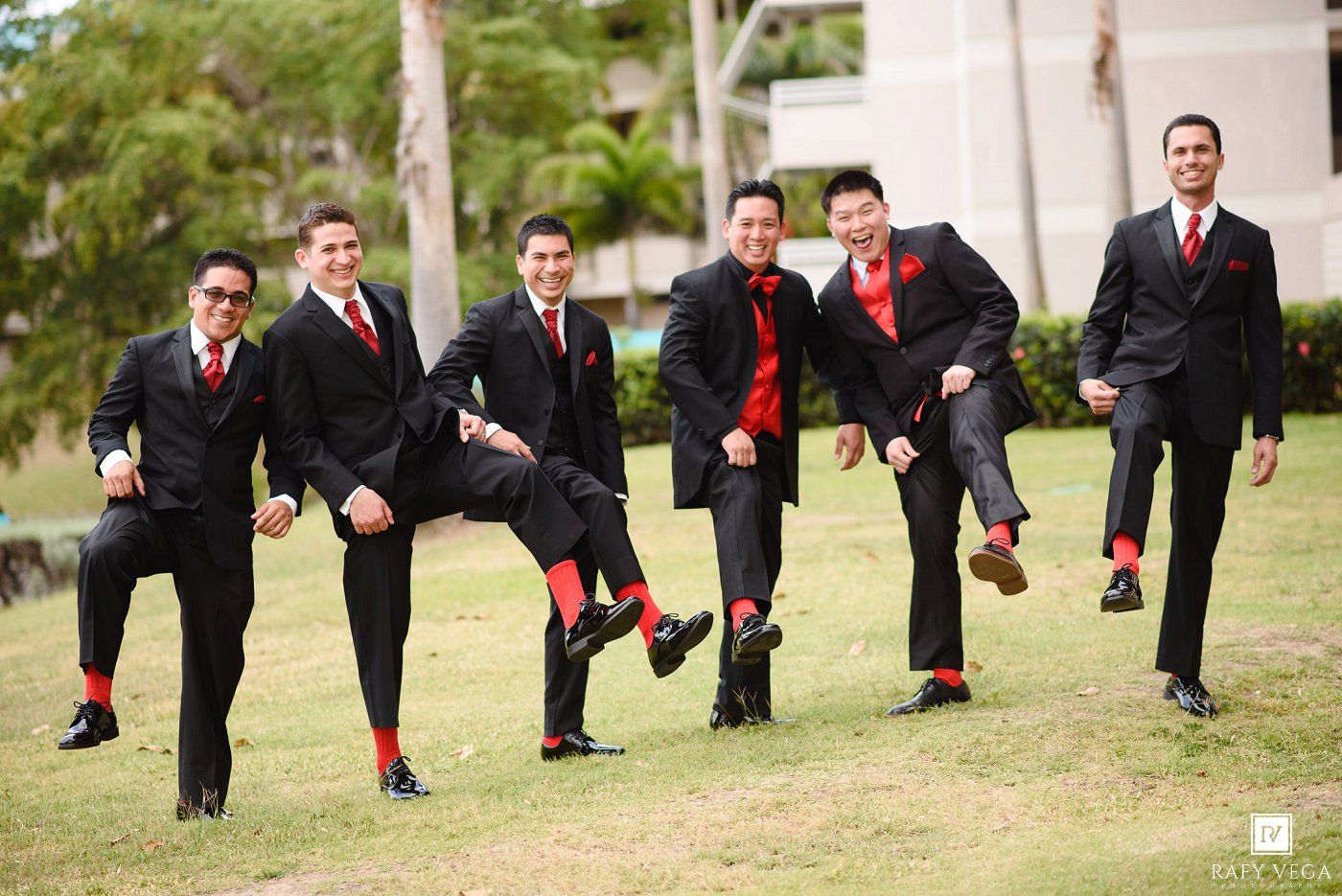 Six groomsmen in matching black suits with red ties and socks strike a fun pose on green grass.