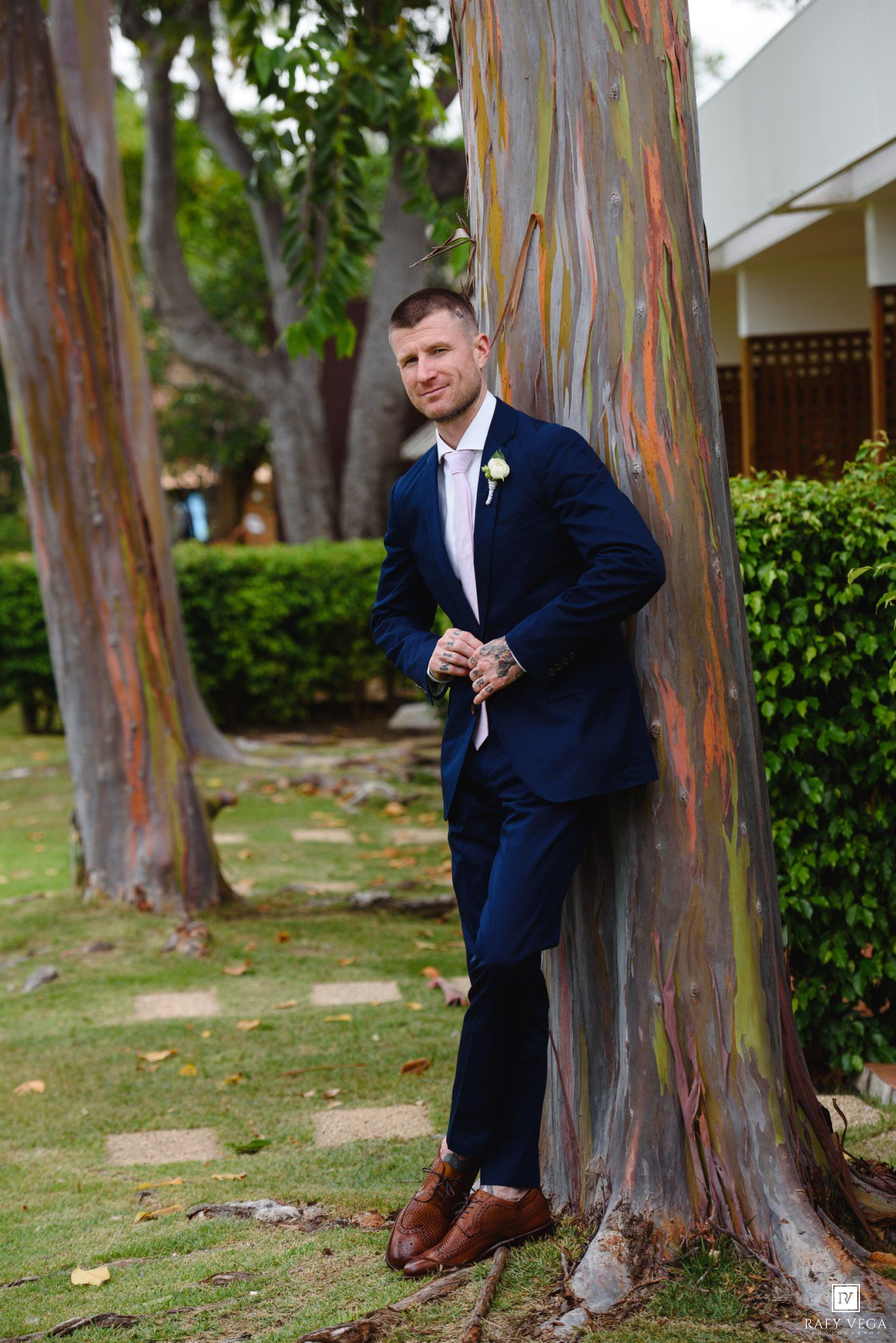 Groom in navy blue suit with boutonniere leans against colorful rainbow eucalyptus tree at outdoor wedding venue.