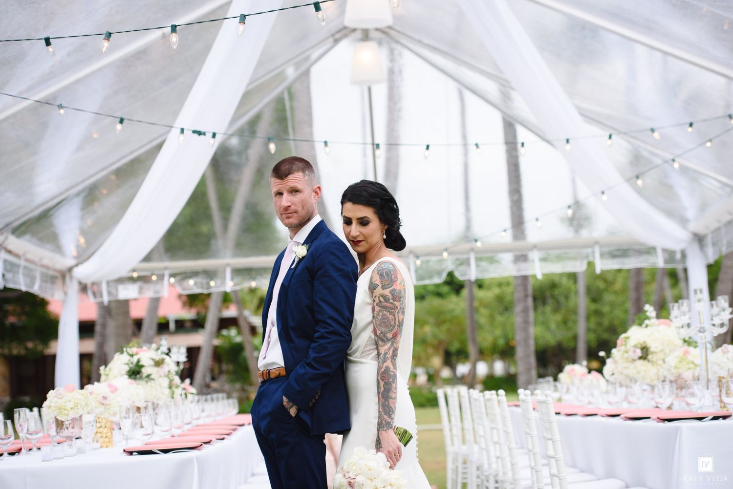 Bride and groom pose elegantly under a draped tent at a romantic outdoor wedding reception with floral decor.