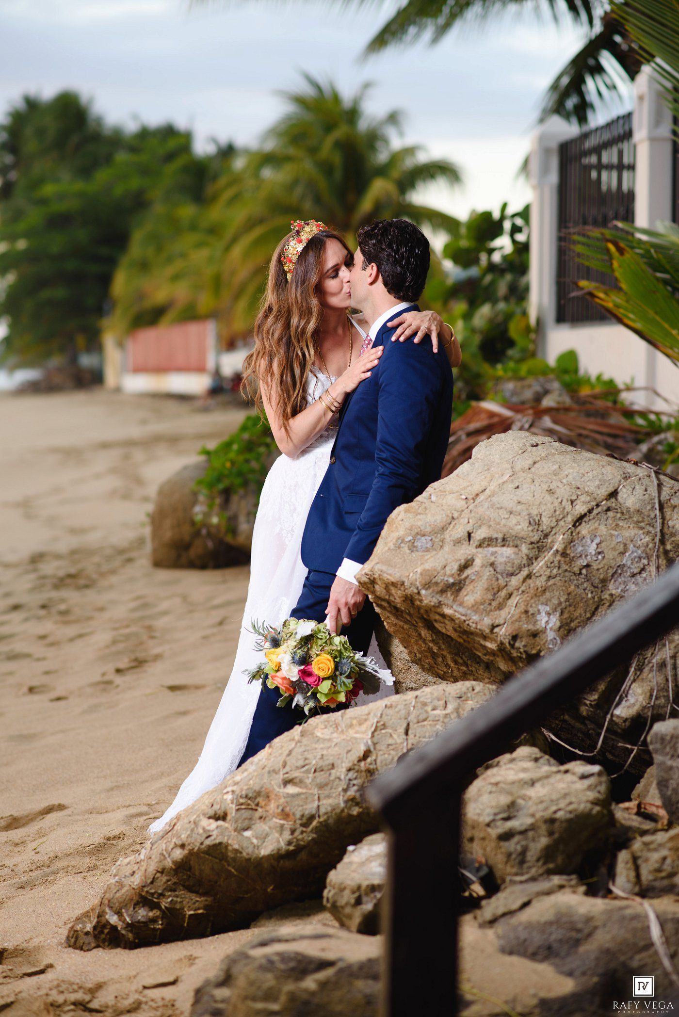 Bride and groom share a romantic kiss on a tropical beach, with palm trees and sand in the background.