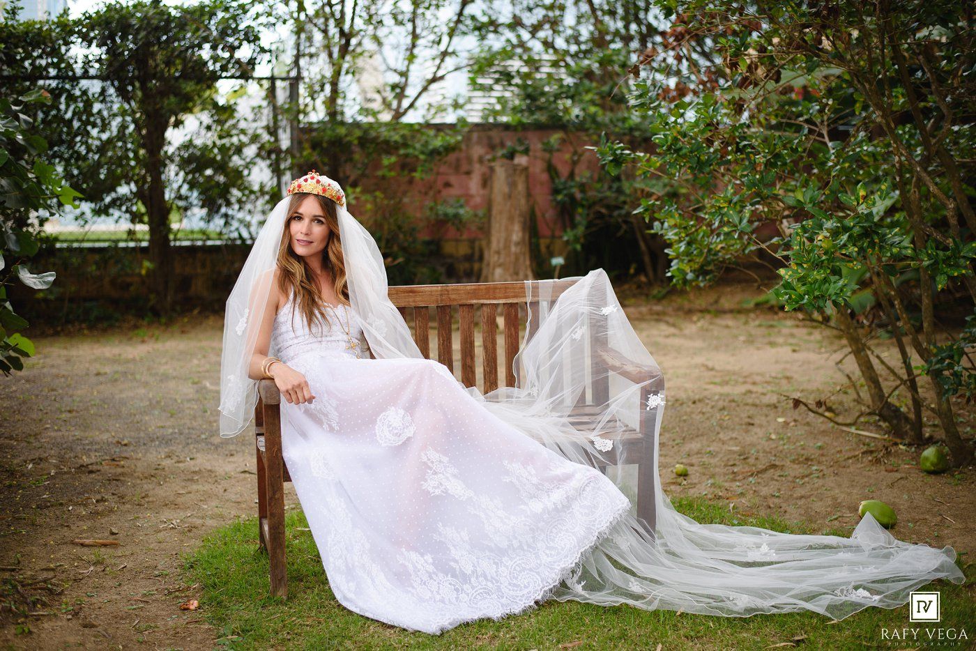 Bride in white lace gown and floral crown sits gracefully on a wooden bench in a garden setting.