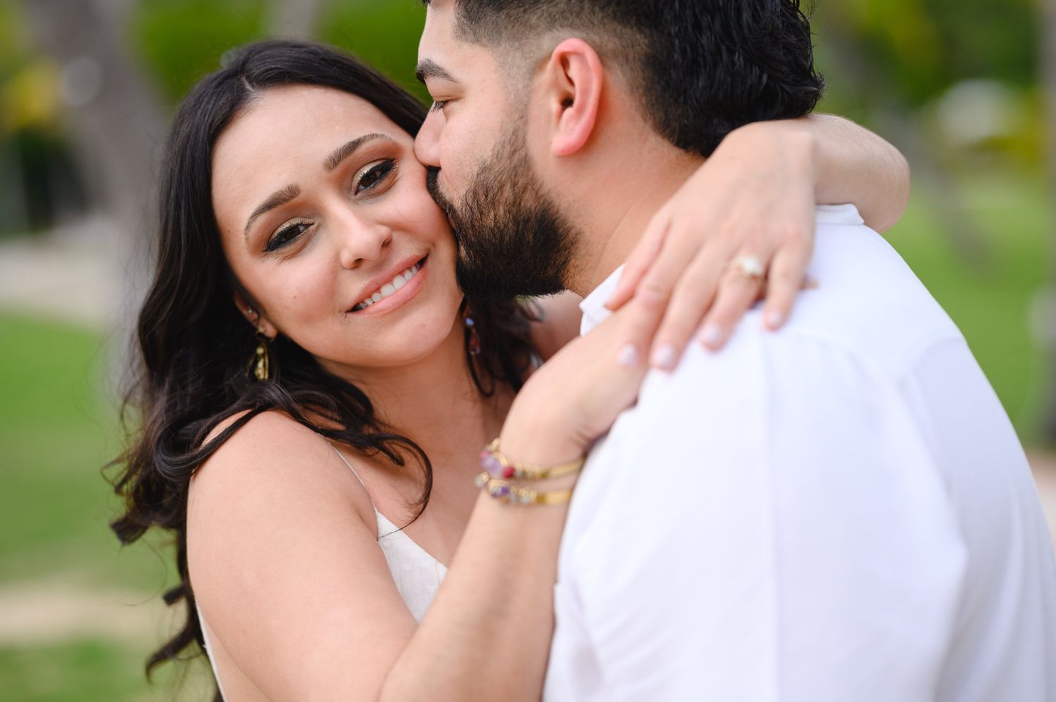 A smiling bride embraces her groom outdoors, wearing a white dress and gold jewelry in a romantic couples portrait.