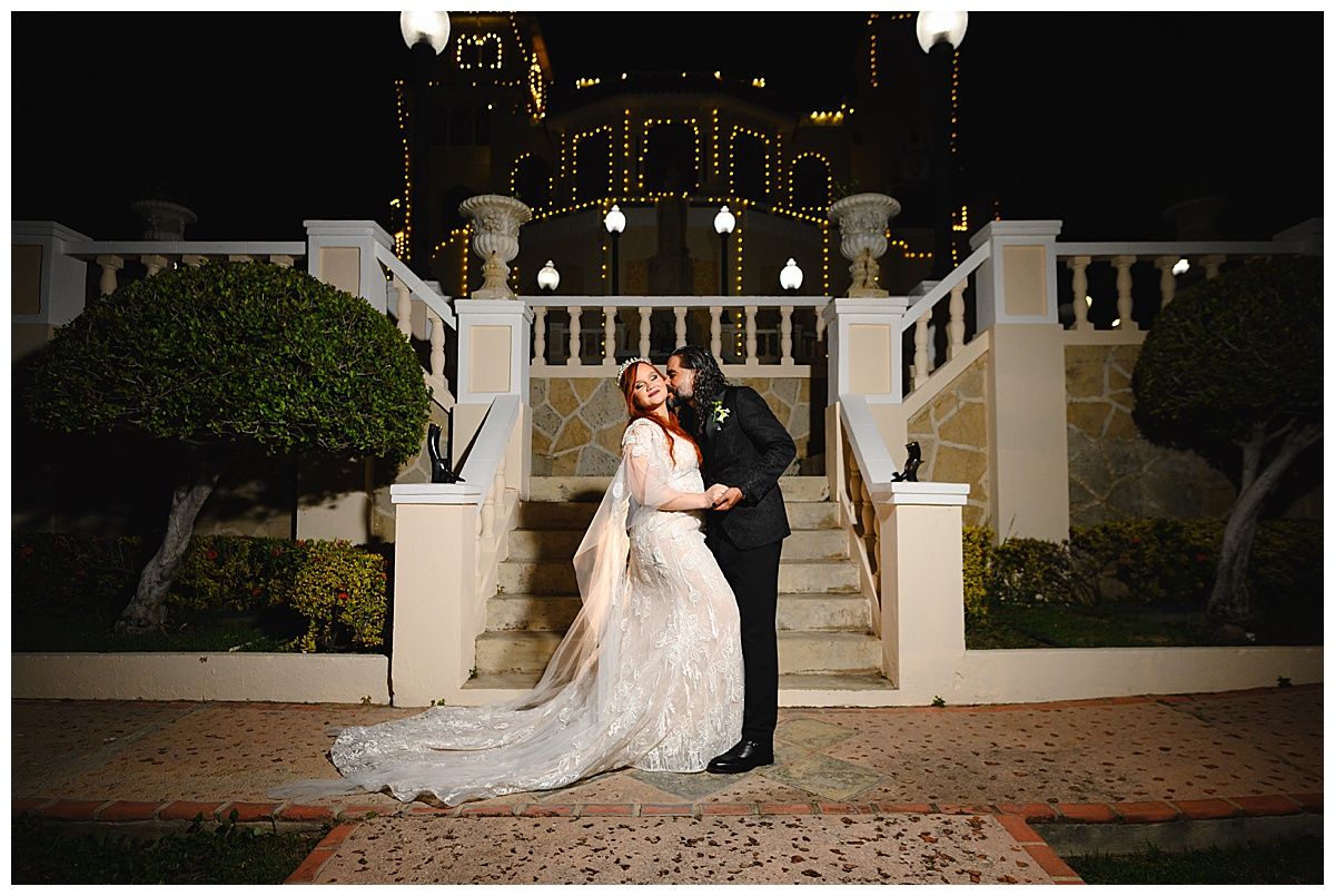 Bride and groom share romantic kiss outside illuminated venue staircase at night.