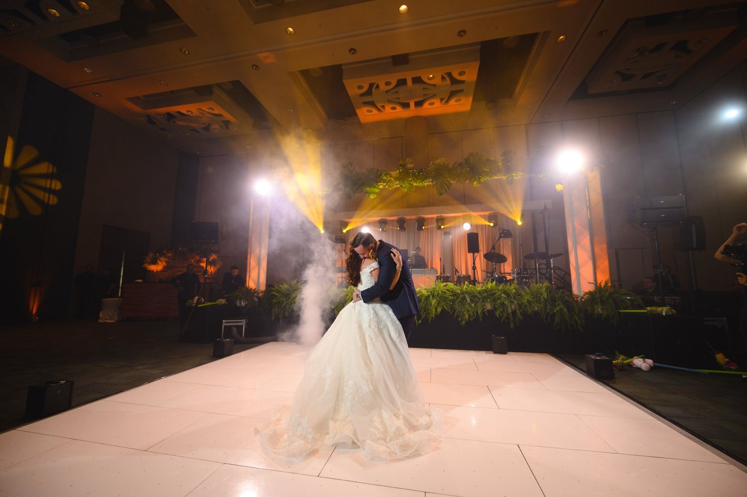 Bride and groom share first dance on white dance floor with dramatic fog and warm stage lighting at wedding reception.