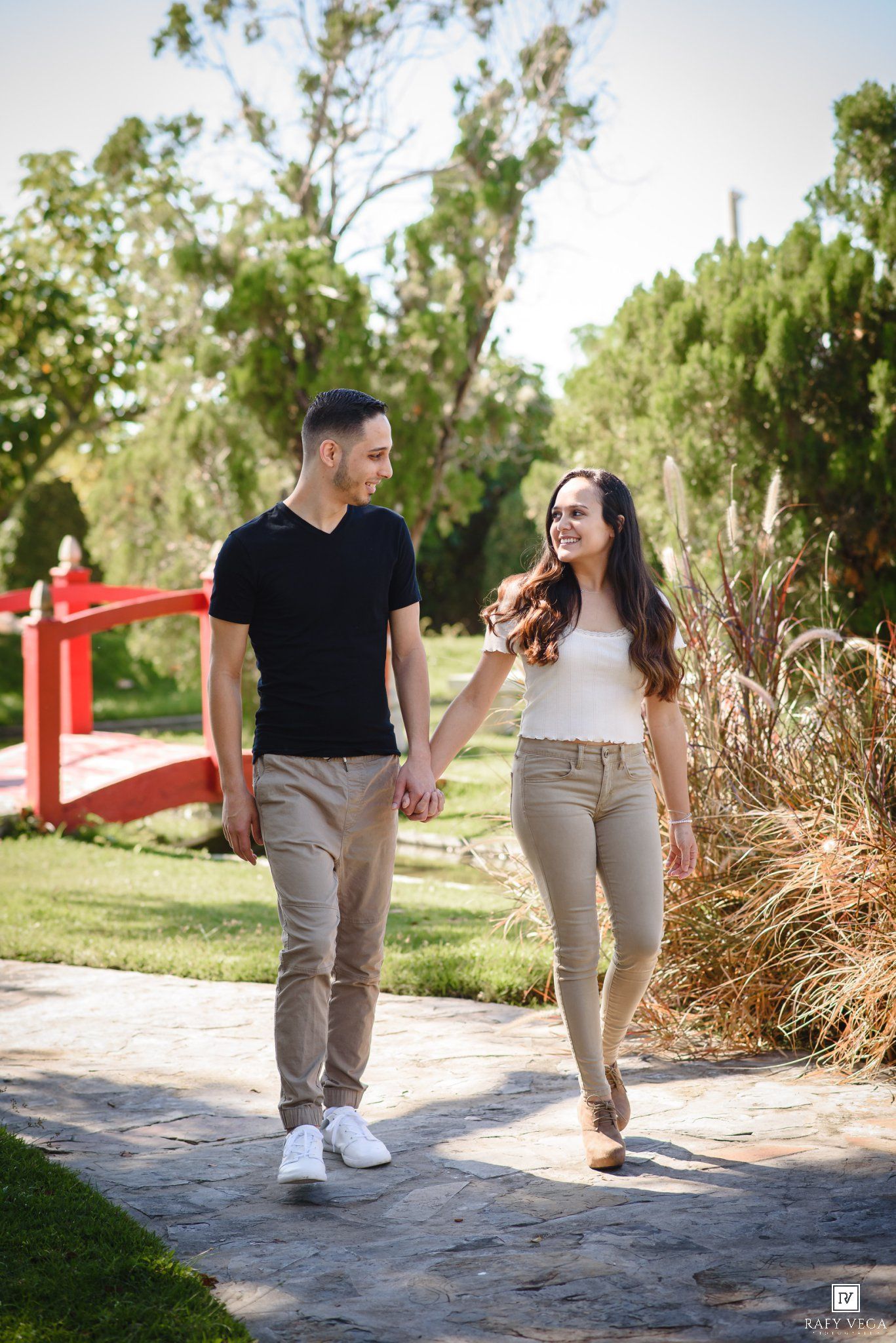 A smiling couple holds hands while walking along a stone path in a lush green park with a red bridge.