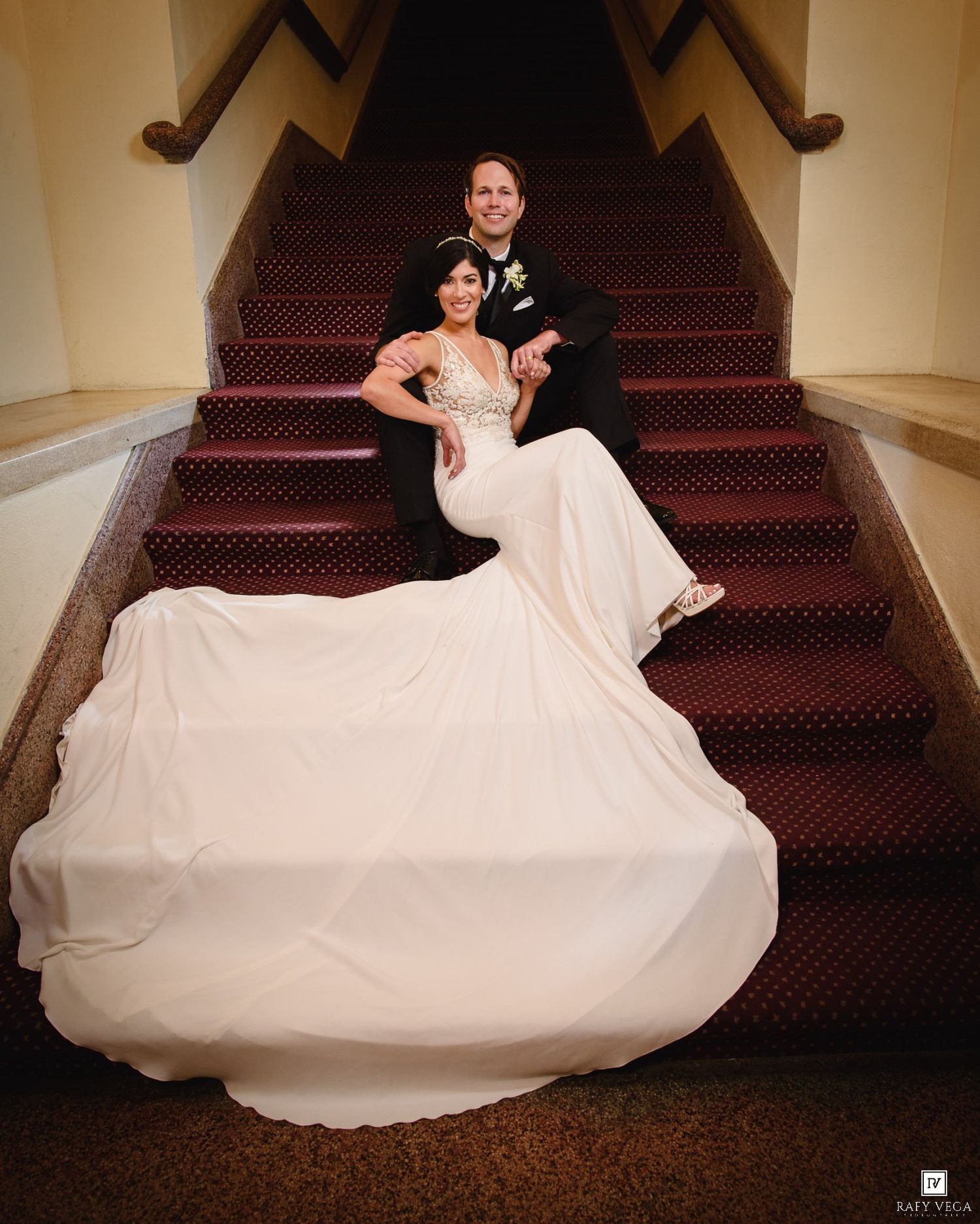 Bride in flowing white gown and groom in black tuxedo pose elegantly on red carpeted staircase for wedding portrait.