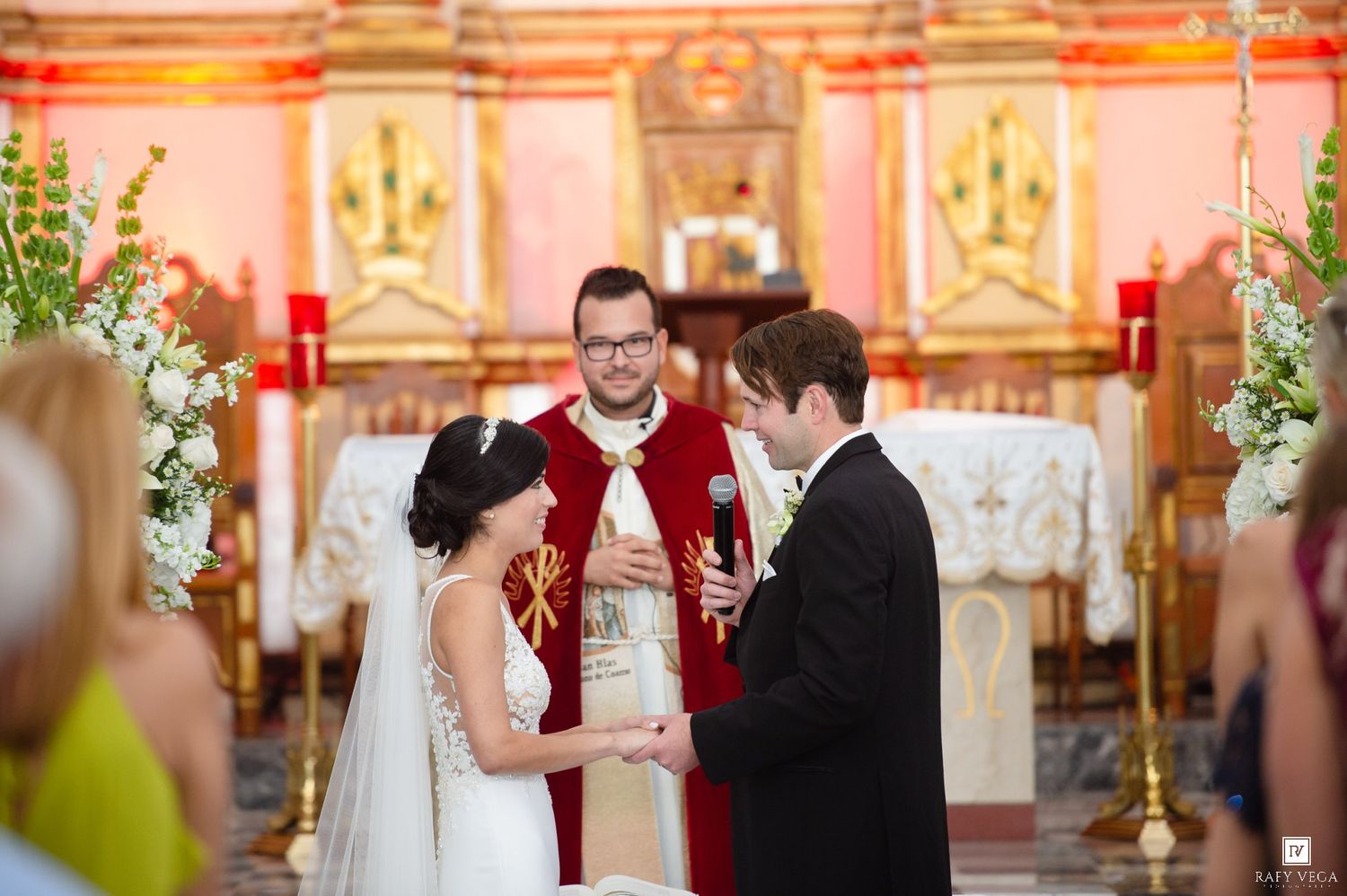 Priest officiating wedding ceremony as bride and groom exchange vows at decorated Catholic church altar.