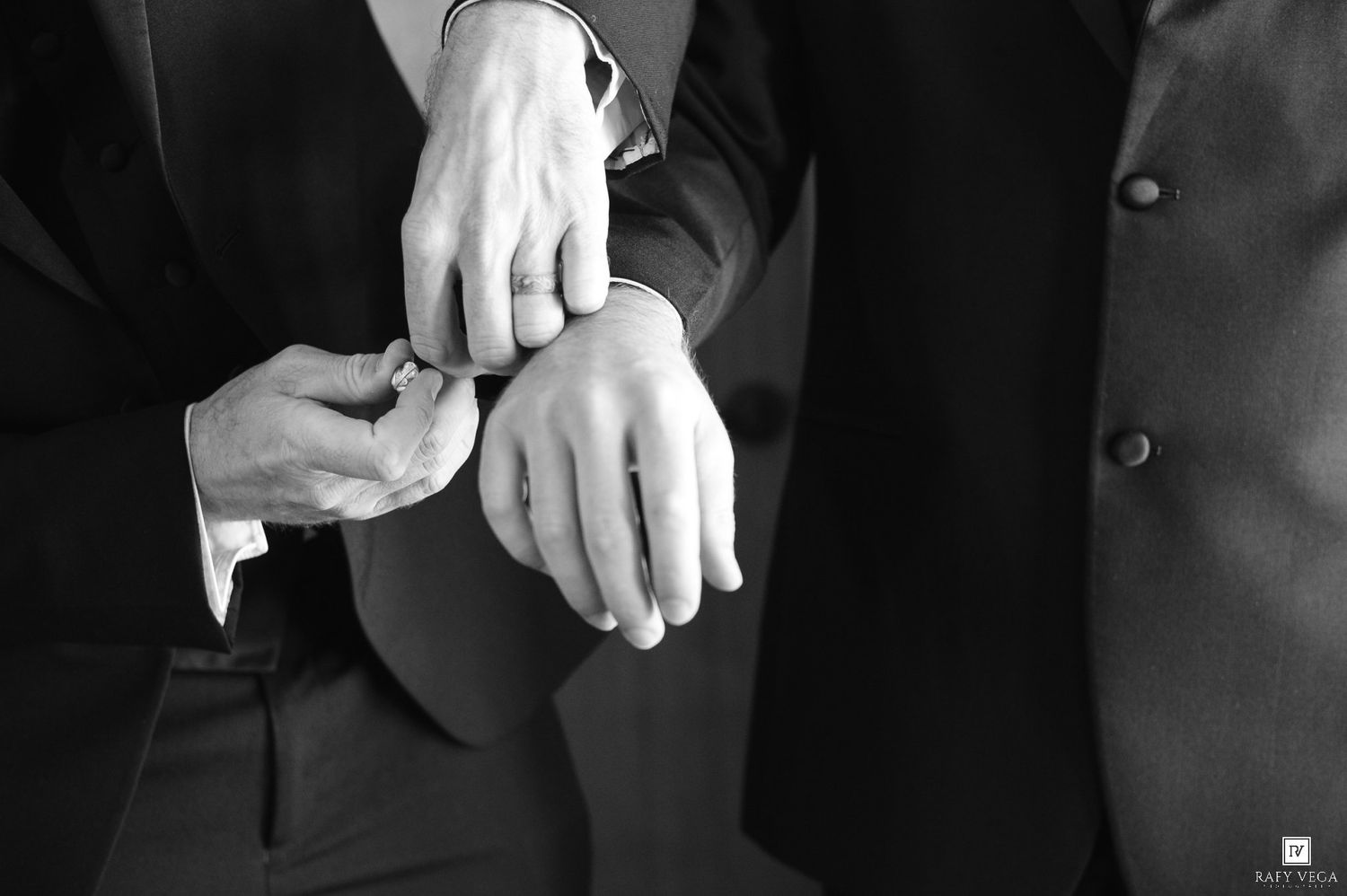 Black and white photo of two men in suits, one fastening the other's cufflink, elegant wedding preparation detail.
