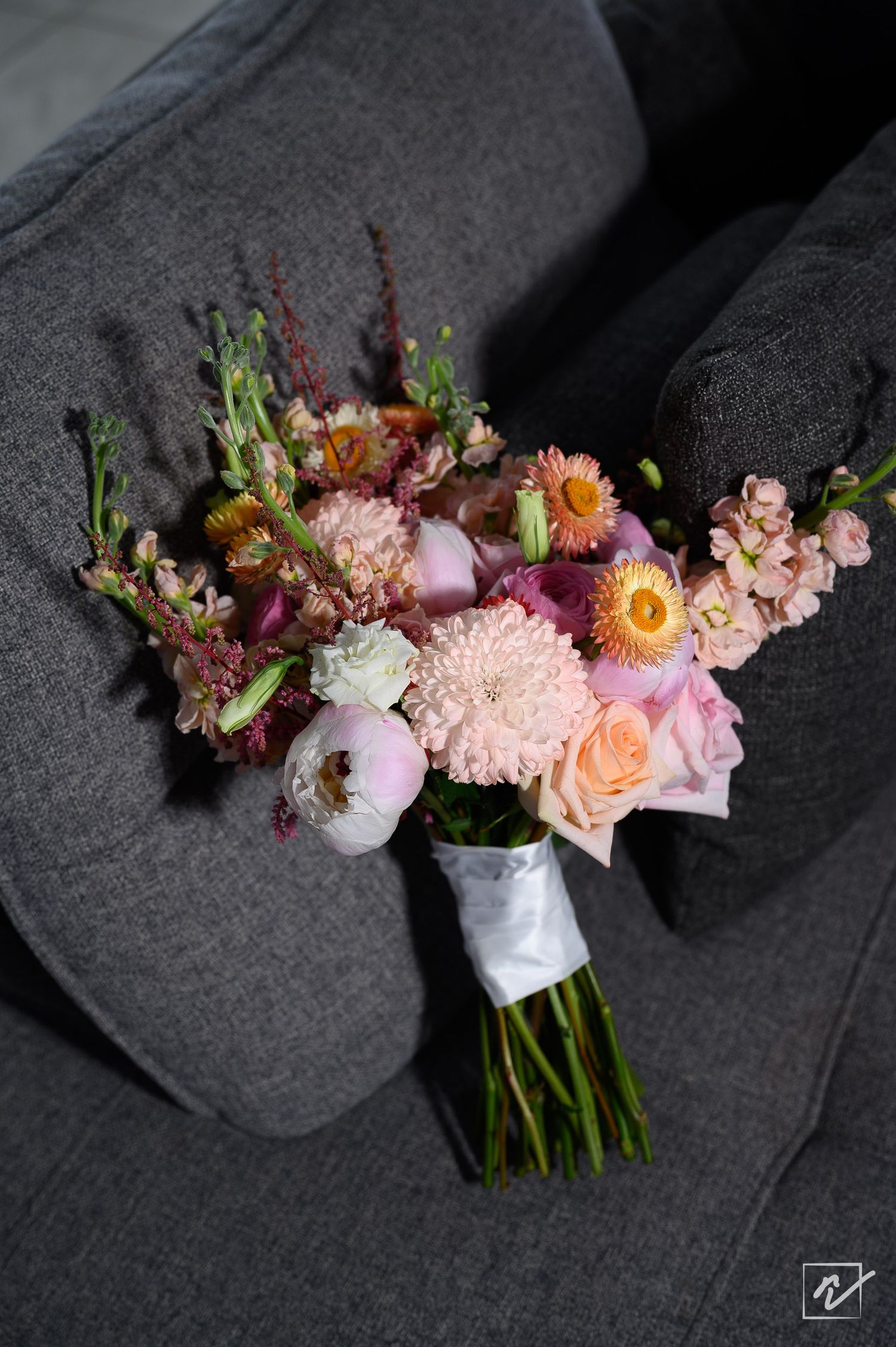 Pink and peach wedding bouquet with dahlias, ranunculus and stock flowers wrapped in white ribbon, resting on a gray upholstered chair.
