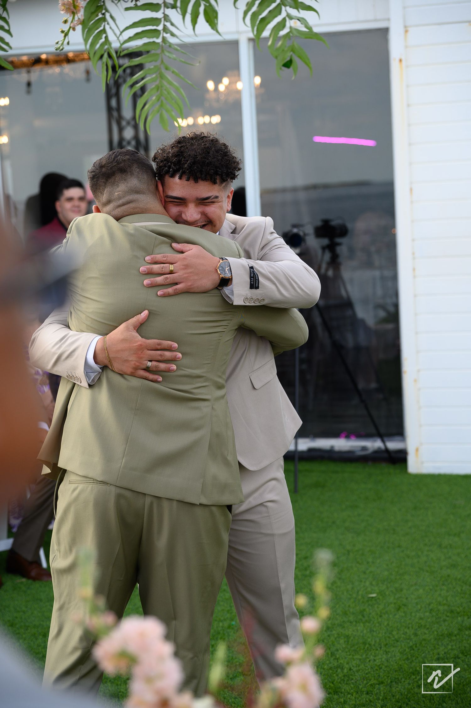 Two men in sage green and tan wedding suits share a warm hug on an outdoor lawn, with pink flowers and a glass venue behind them.