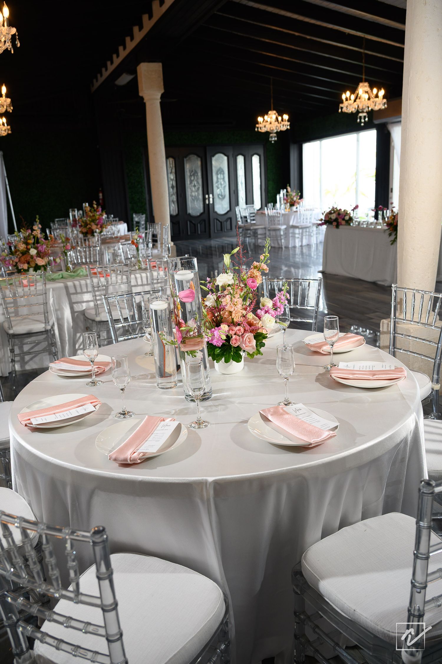 Elegant wedding reception table with white linen, pink napkins, floral centerpiece, and clear Chiavari chairs under crystal chandeliers.