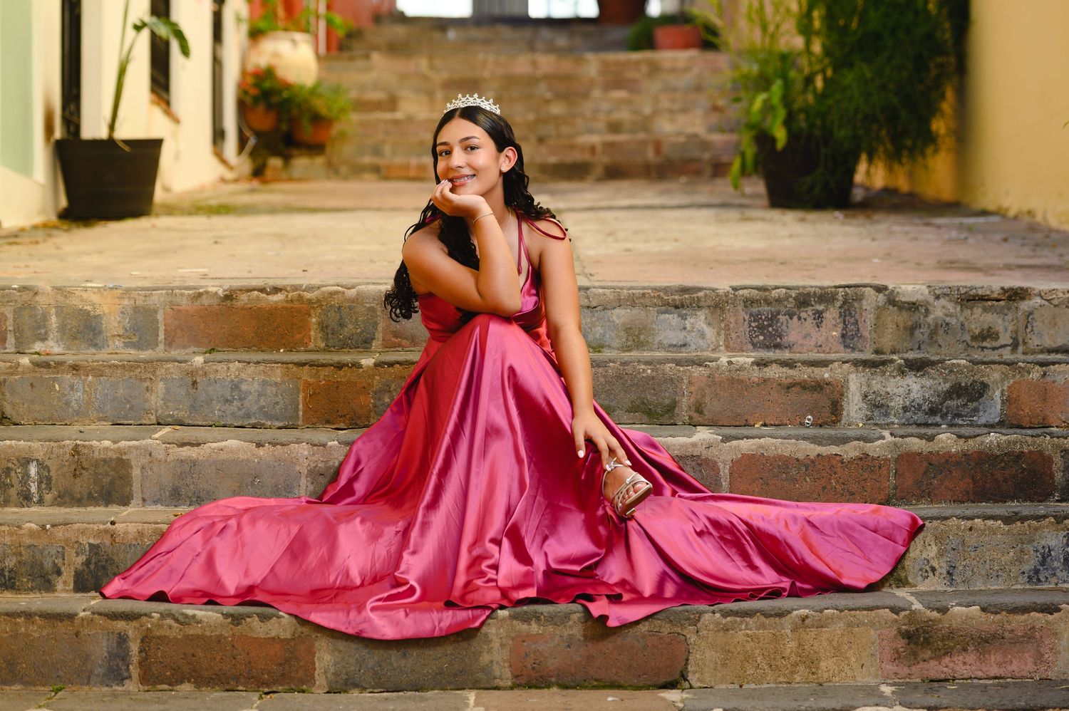 Quinceañera in a pink satin ballgown and tiara poses gracefully on stone steps outdoors.