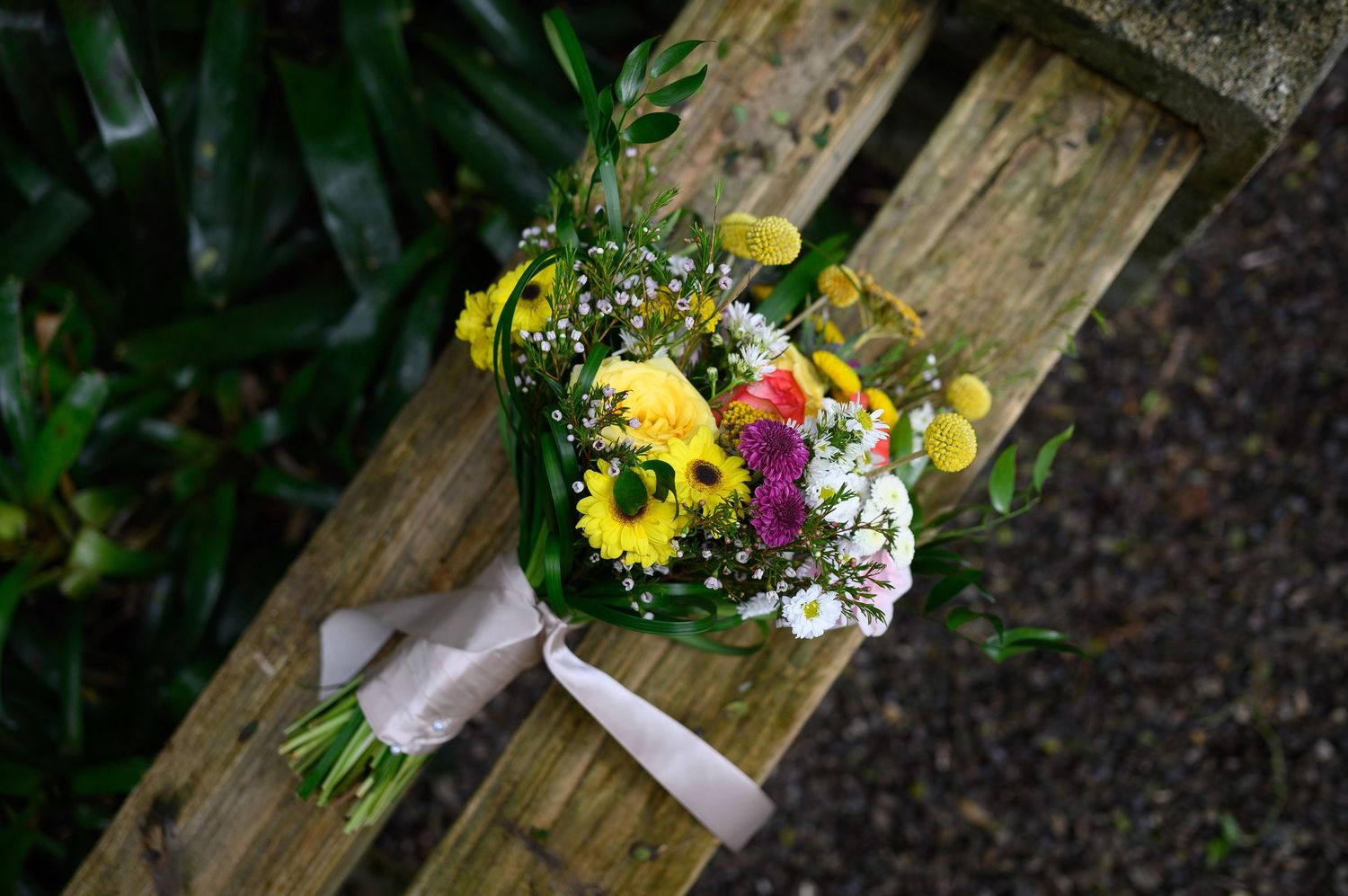 Colorful wildflower bouquet with yellow gerberas, white baby's breath, and pink blooms tied with ribbon on a wooden bench.
