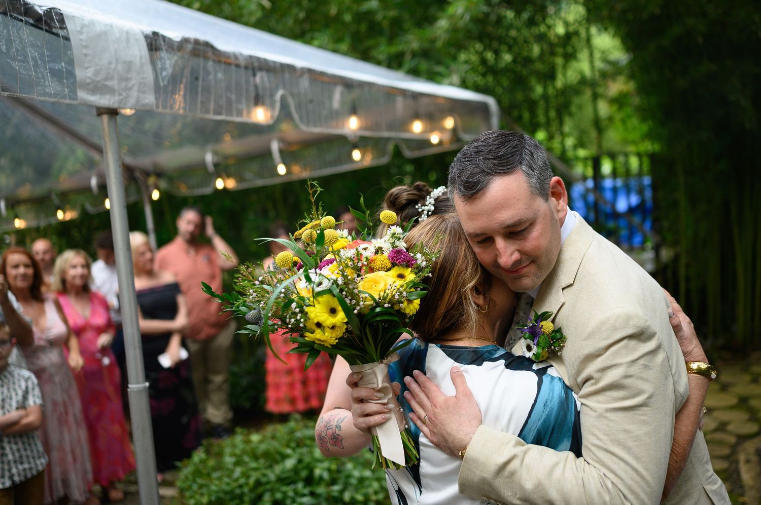 Bride and groom share a heartfelt hug at outdoor wedding reception under tent with floral bouquet.