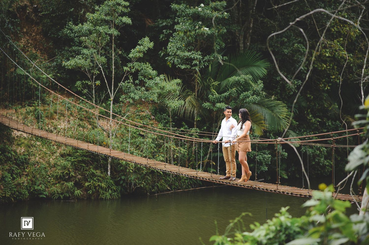 Couple standing on a rustic rope suspension bridge surrounded by lush green tropical forest trees.
