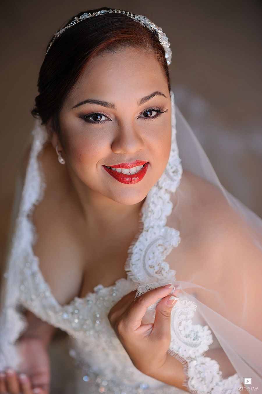 Smiling bride in beaded white gown and lace veil with crystal headband and red lips on wedding day.