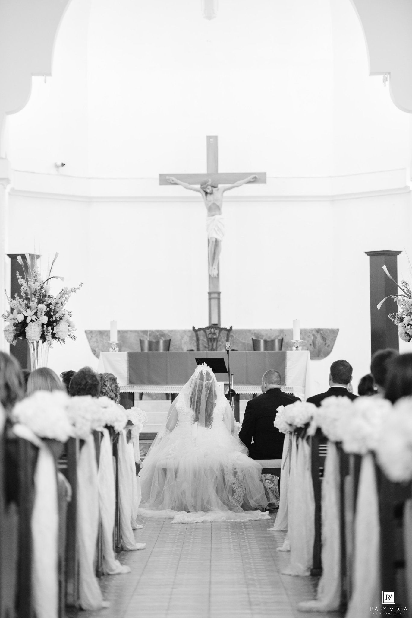 Black and white photo of a bride in a veil walking down the church aisle toward a crucifix during a wedding ceremony.
