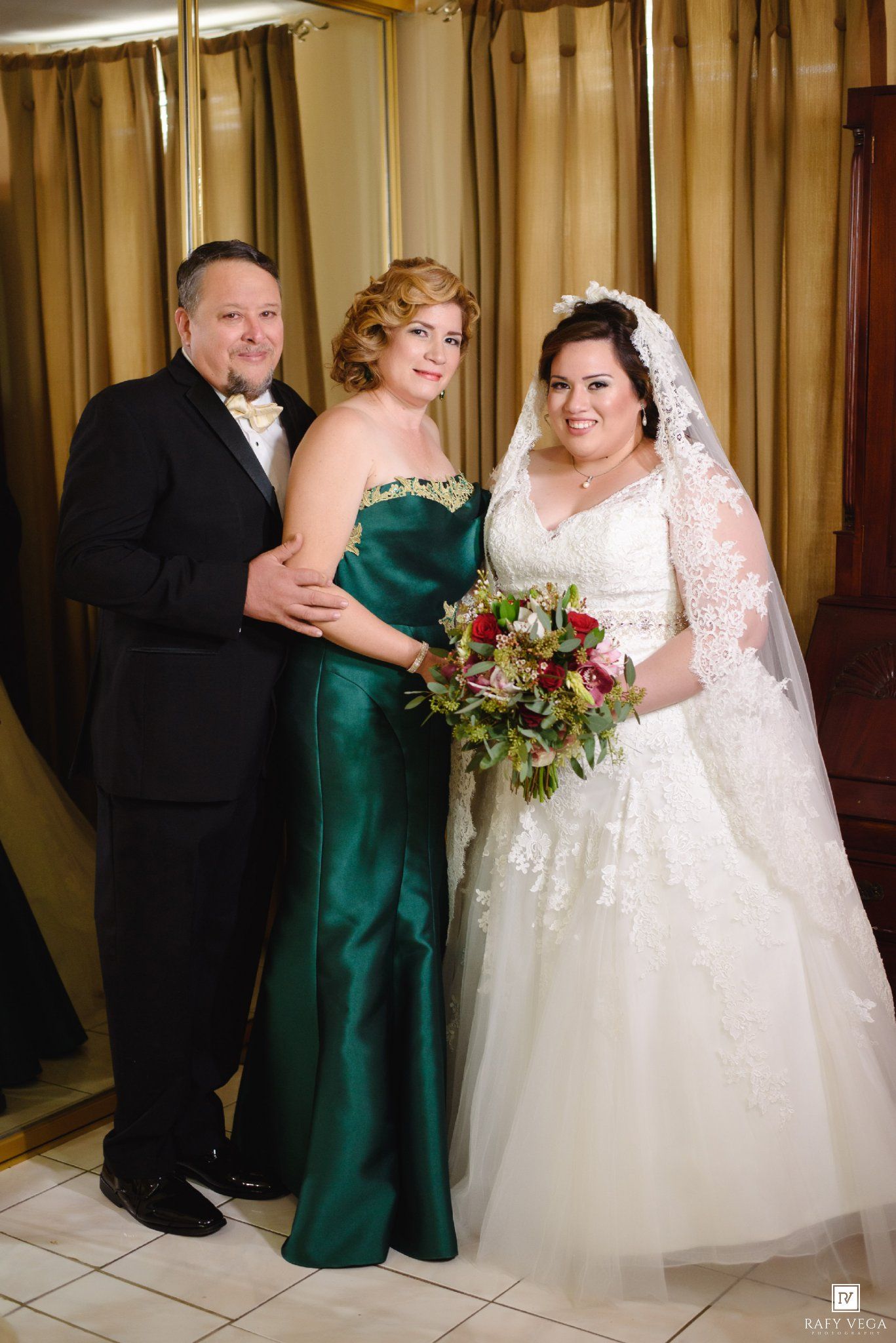 Bride in white lace gown with veil holds bouquet beside man in tuxedo and woman in emerald green gown.