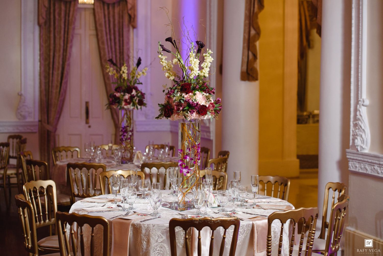 Elegant wedding reception hall with gold Chiavari chairs, round tables, and tall floral centerpieces in a grand ballroom.