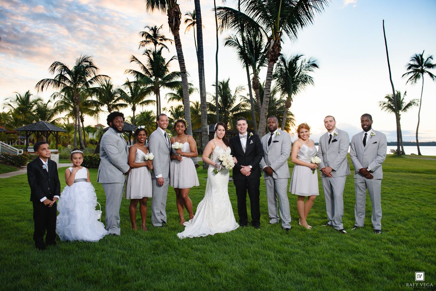 Wedding party poses on tropical lawn with palm trees at sunset, bride and groom centered among groomsmen and bridesmaids.