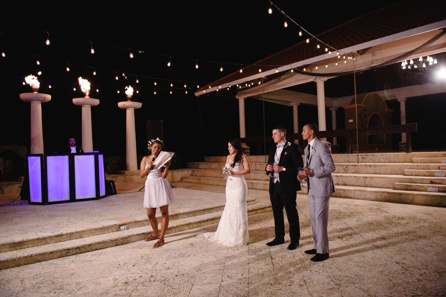 Outdoor wedding ceremony at night with string lights, couple and officiant standing on stone patio near torches.
