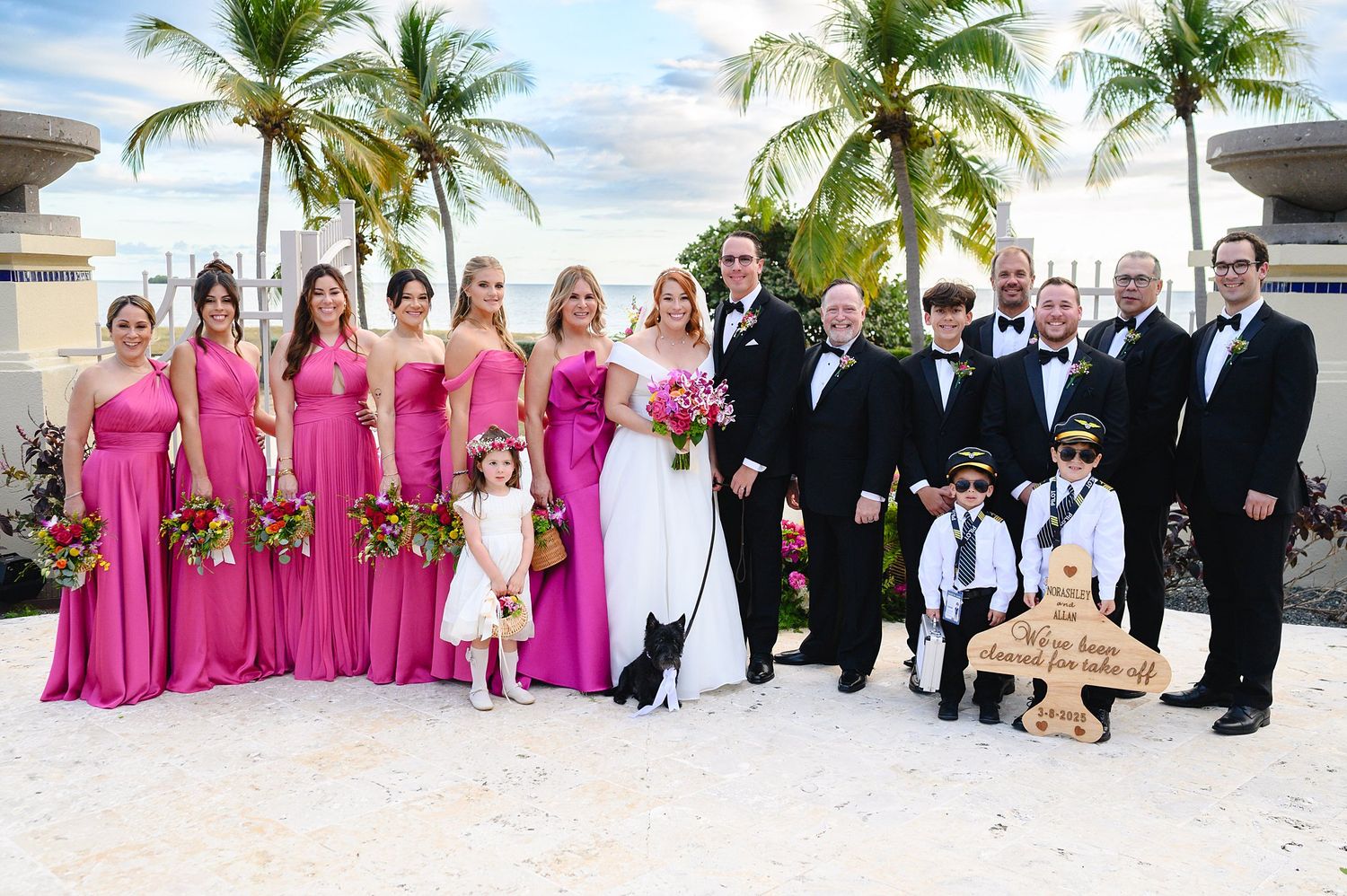 Wedding party poses on beach with bridesmaids in pink gowns, groomsmen in tuxedos, and couple in white and black attire.
