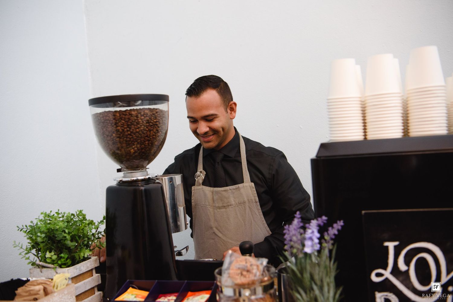 Smiling barista in apron preparing coffee at a professional espresso station with coffee grinder and stacked cups.
