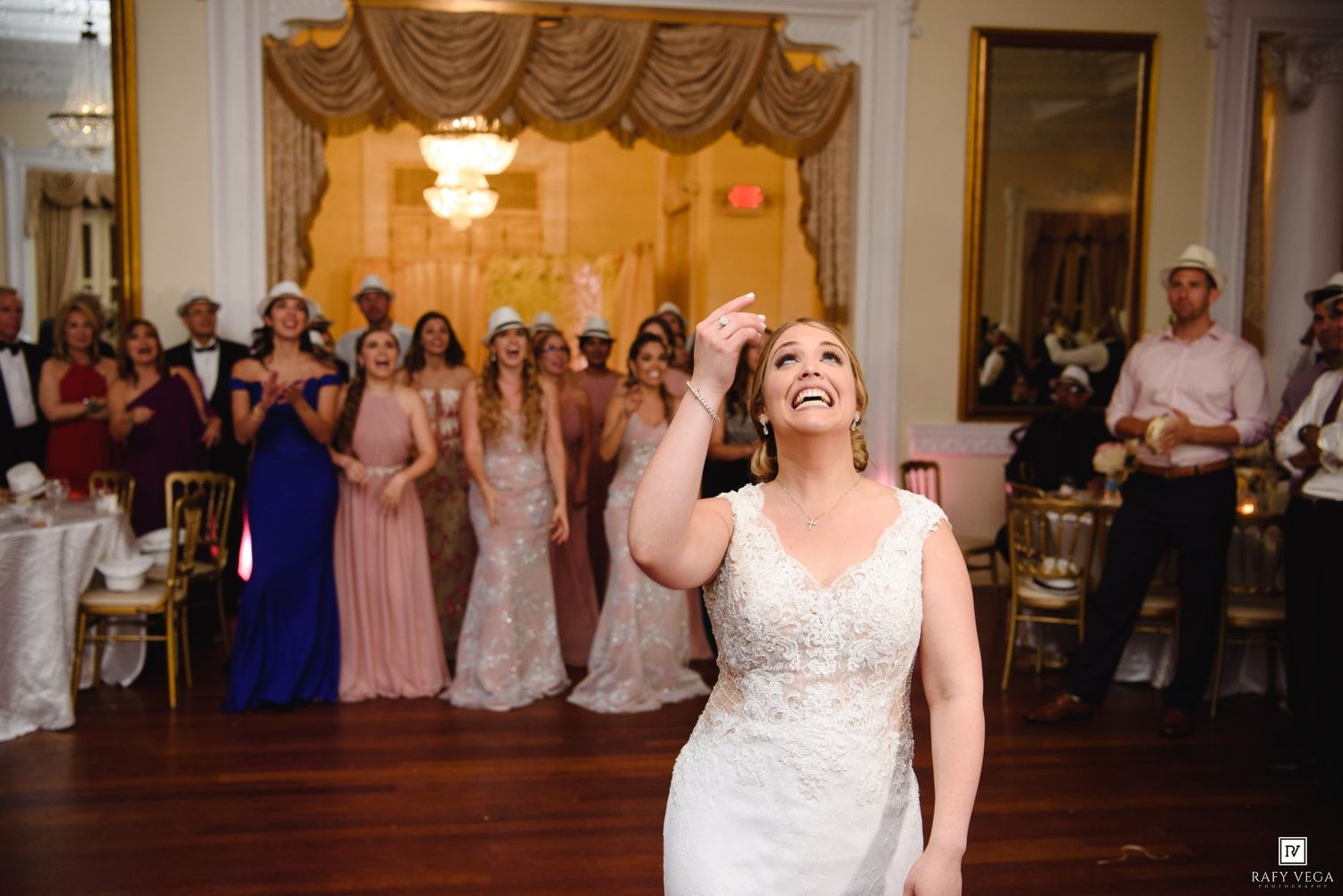 Bride in white gown laughing while tossing bouquet at elegant wedding reception with bridesmaids in pink dresses.