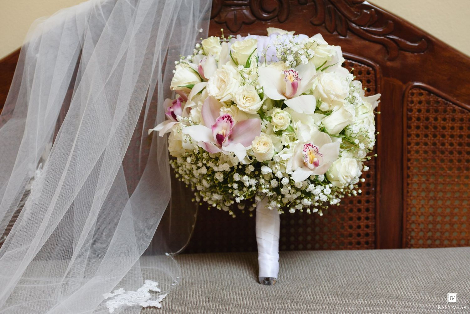 Elegant bridal bouquet of white roses, pink orchids, and baby's breath beside a flowing wedding veil on a wooden chair.