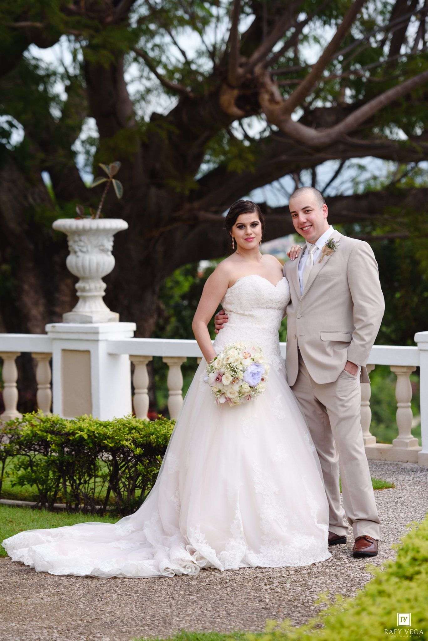 Bride in white gown holding bouquet stands with groom in tan suit at elegant outdoor garden wedding venue.