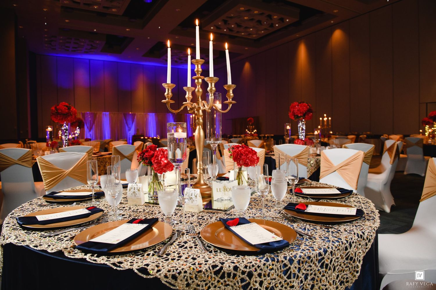 Elegant wedding reception table with gold candelabra, red roses, lace tablecloth, and gold charger plates in a ballroom.