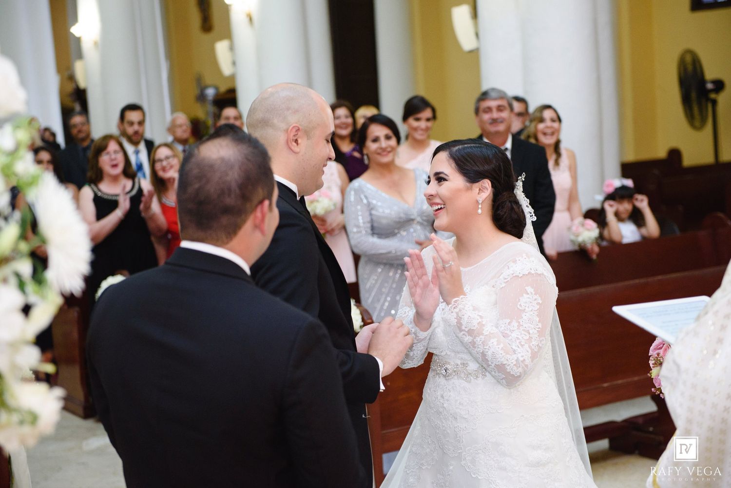 Bride in lace gown smiling joyfully at groom during church wedding ceremony with guests seated in pews.